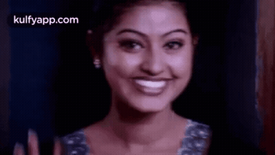 A Woman Is Smiling And Waving Her Hand In Front Of A Dark Background . GIF