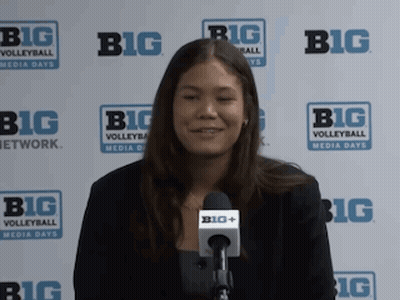 A Woman Is Smiling In Front Of A Big Volleyball Sign GIF