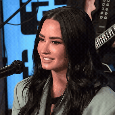 A Woman Is Smiling In Front Of A Microphone With A Guitar In The Background GIF