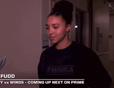 A Woman Is Smiling In Front Of A Sign That Says Fuddy Vs Wings Coming Up Next On Prime GIF