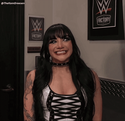 A Woman Is Smiling In Front Of A Sign That Says The Wrestling Factory GIF