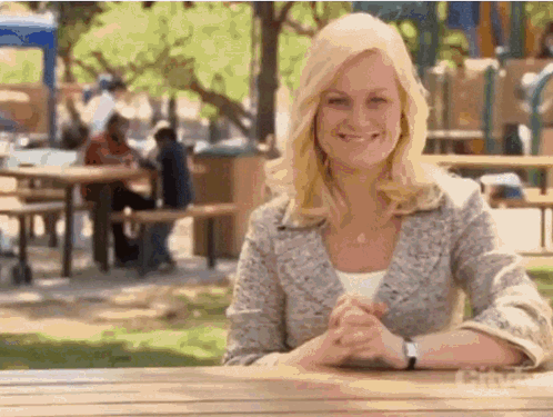 A Woman Is Smiling While Sitting At A Picnic Table In A Park . GIF