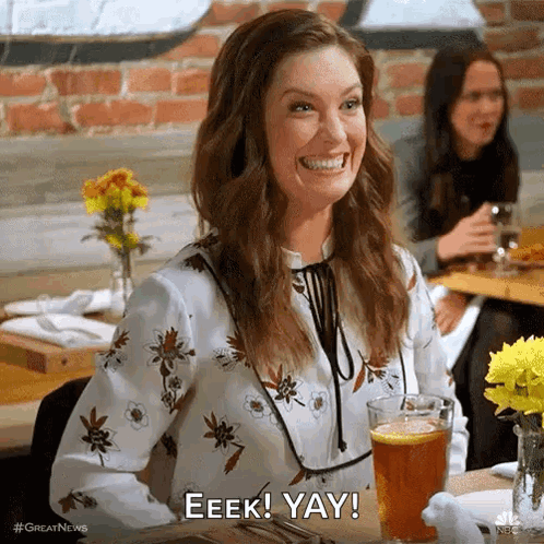 A Woman Is Smiling While Sitting At A Table With A Glass Of Beer And Flowers . GIF