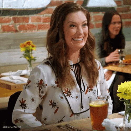 A Woman Is Smiling While Sitting At A Table With A Glass Of Beer And Flowers . GIF