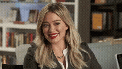 A Woman Is Smiling While Sitting In Front Of A Bookshelf . GIF