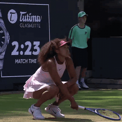 A Woman Is Squatting Down With A Tennis Racquet In Front Of An Advertisement For Tutima Glashutte GIF
