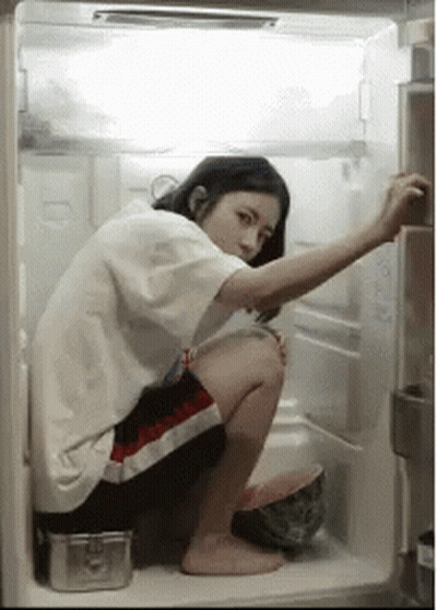 A Woman Is Squatting In An Open Refrigerator Looking For Something To Eat . GIF