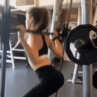 A Woman Is Squatting With A Barbell On Her Shoulders In A Gym . GIF