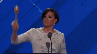A Woman Is Standing At A Podium With A Microphone And Holding A Maracas In Her Hand . GIF