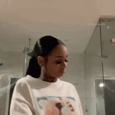 A Woman Is Standing In A Bathroom Wearing A White T-Shirt And A Ponytail . GIF