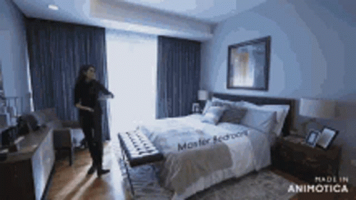 A Woman Is Standing In A Bedroom With A Large Bed And A Bench . GIF