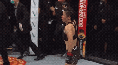 A Woman Is Standing In A Boxing Ring With A Body Armor Drink In The Background . GIF
