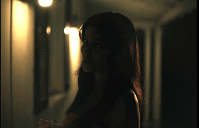 A Woman Is Standing In A Dark Hallway With A Light Shining On Her GIF