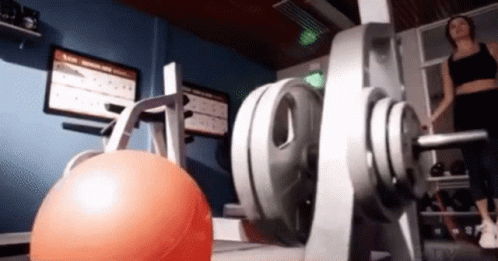 A Woman Is Standing In A Gym Next To A Dumbbell GIF
