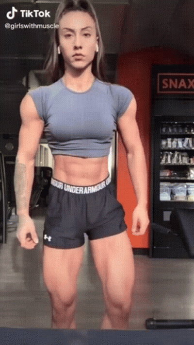 A Woman Is Standing In A Gym Wearing Under Armour Shorts And A Blue Crop Top . GIF