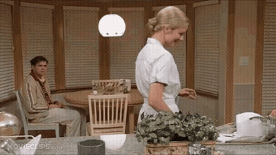 A Woman Is Standing In A Kitchen With A Man Sitting At A Table . GIF
