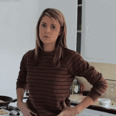 A Woman Is Standing In A Kitchen With Her Hands On Her Hips . She Is Wearing A Brown Striped Shirt . GIF