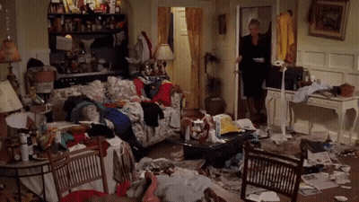 A Woman Is Standing In A Messy Living Room Filled With Lots Of Clothes And Papers . GIF