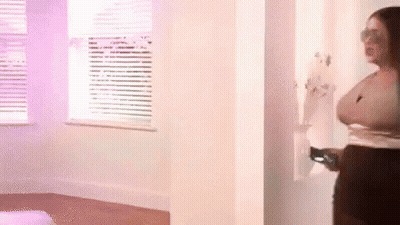 A Woman Is Standing In A Room Next To A Window Holding A Remote Control . GIF