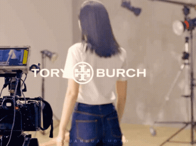 A Woman Is Standing In Front Of A Camera With Tory Burch Written On It GIF