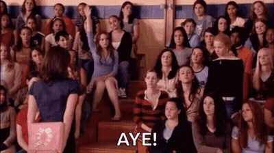 A Woman Is Standing In Front Of A Crowd Of People And Raising Her Hand To Answer A Question . GIF