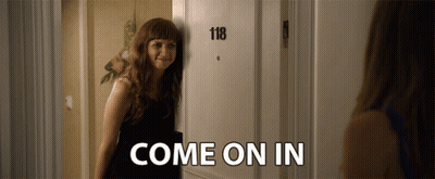 A Woman Is Standing In Front Of A Door That Says 118 And Says Come On In GIF