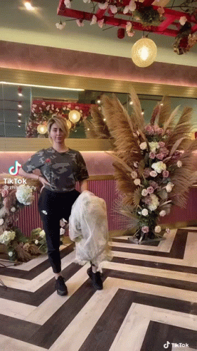A Woman Is Standing In Front Of A Floral Arrangement In A Room With A Striped Floor . GIF