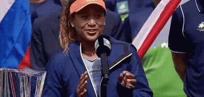 A Woman Is Standing In Front Of A Microphone And Smiling While Wearing An Orange Hat . GIF