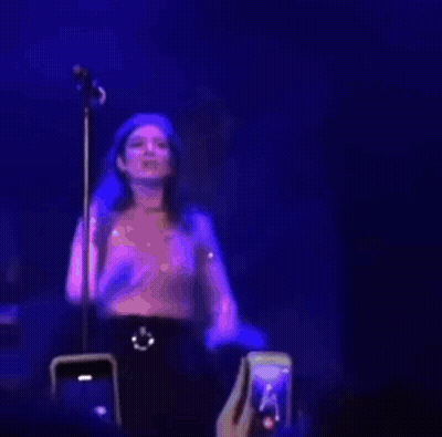 A Woman Is Standing In Front Of A Microphone On A Stage While A Fan Takes A Picture Of Her . GIF