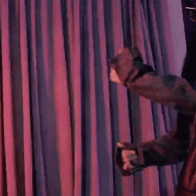A Woman Is Standing In Front Of A Purple Curtain Holding A Microphone And Blowing A Kiss . GIF