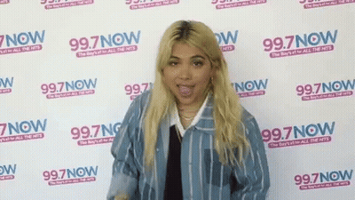 A Woman Is Standing In Front Of A Wall That Says 99.7 Now GIF