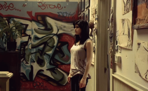 A Woman Is Standing In Front Of A Wall With Graffiti On It That Says " Steve " GIF