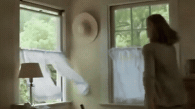 A Woman Is Standing In Front Of A Window With A Hat On The Wall GIF