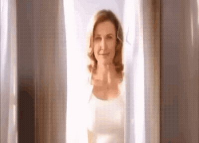 A Woman Is Standing In Front Of A Window With White Curtains And Smiling . GIF