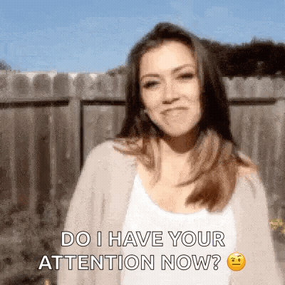 A Woman Is Standing In Front Of A Wooden Fence And Says Do I Have Your Attention Now . GIF