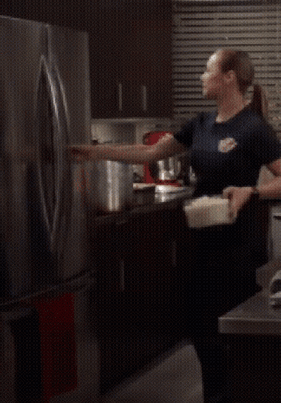 A Woman Is Standing In Front Of An Open Refrigerator Holding A Container GIF