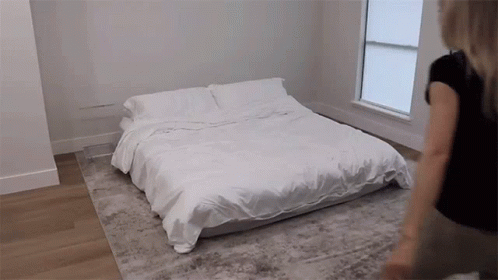 A Woman Is Standing Next To A Bed With White Sheets And Pillows GIF