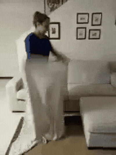 A Woman Is Standing Next To A White Couch In A Living Room GIF