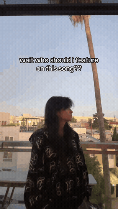 A Woman Is Standing On A Balcony With The Caption Wait Who Should I Feature On This Song ? GIF