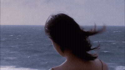 A Woman Is Standing On A Beach With Her Hair Blowing In The Wind And Looking At The Ocean . GIF