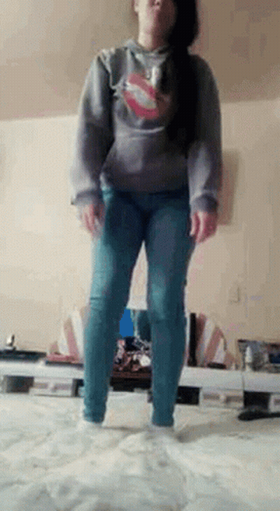 A Woman Is Standing On A Bed Wearing A Hoodie And Jeans . GIF