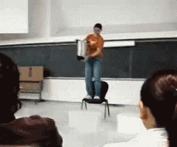 A Woman Is Standing On A Chair In Front Of A Blackboard Holding A Pot GIF