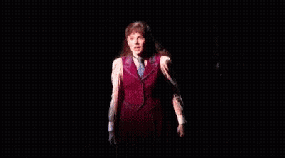 A Woman Is Standing On A Stage In A Dark Room Wearing A Red Vest And Tie . GIF