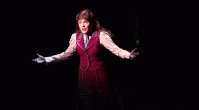A Woman Is Standing On A Stage In A Dark Room Wearing A Red Vest And Tie . GIF