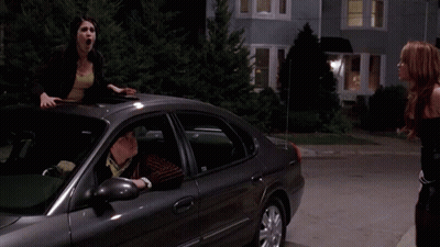 A Woman Is Standing On The Roof Of A Car While Another Woman Looks On With The Words " What ? " Above Her GIF
