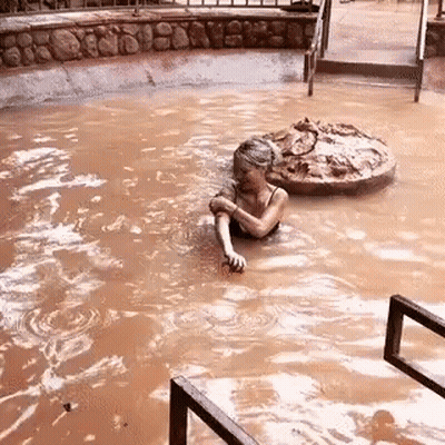 A Woman Is Swimming In A Muddy Pool With A Rock In The Background . GIF