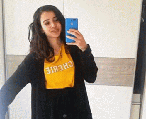 A Woman Is Taking A Selfie In Front Of A Mirror Wearing A Yellow Chebie Shirt GIF