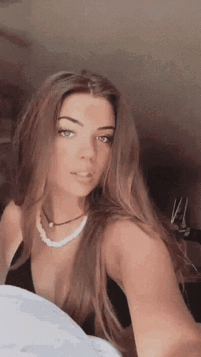 A Woman Is Taking A Selfie With Her Phone While Wearing A Black Top And A Pearl Necklace . GIF
