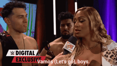 A Woman Is Talking Into A Microphone In Front Of A Sign That Says Vamos ( Let 'S Go ) Boys GIF