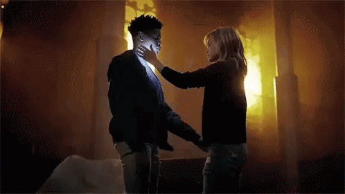 A Woman Is Touching A Man 'S Face In A Room With A Fire In The Background . GIF